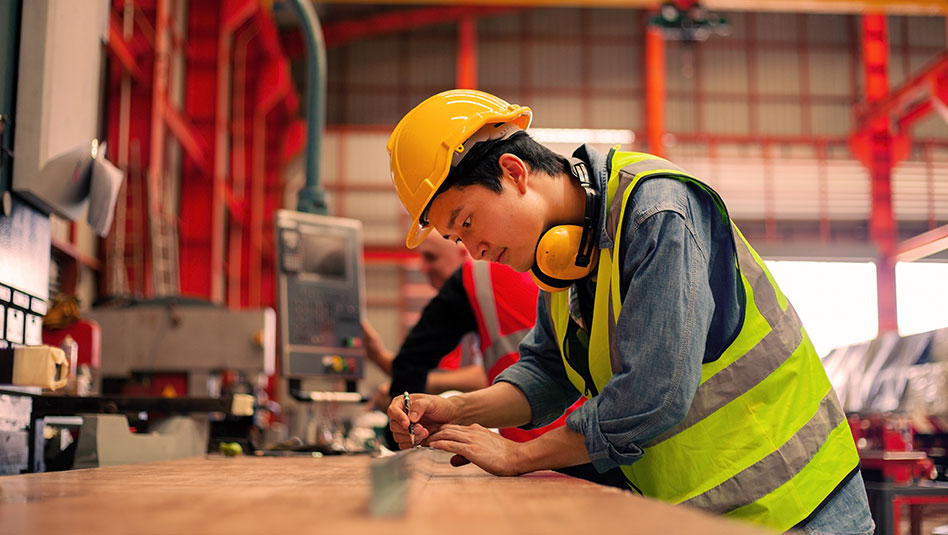 Male factory worker with a hard hat and ear muffs with a pencil in his right hand.