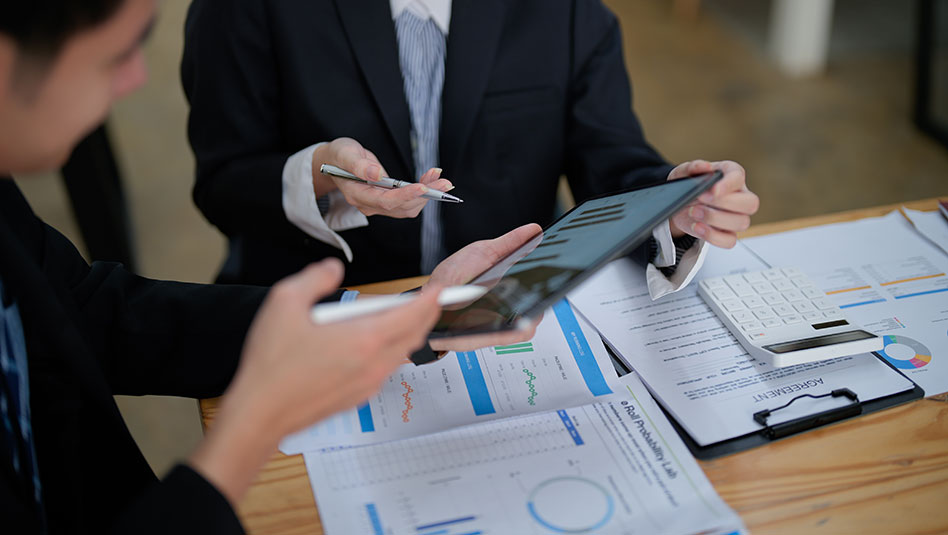 Two persons in suits holding pens in their right hands while talking about some charts.