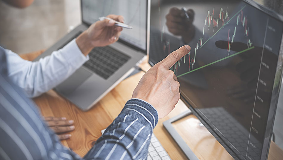 Two office colleagues point to a computer screen showing a candle stick chart with trend lines.