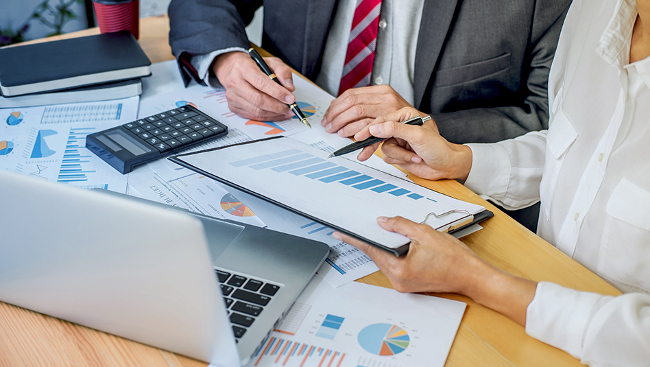 A man and a woman in office attire hold pens as they talk about some charts.