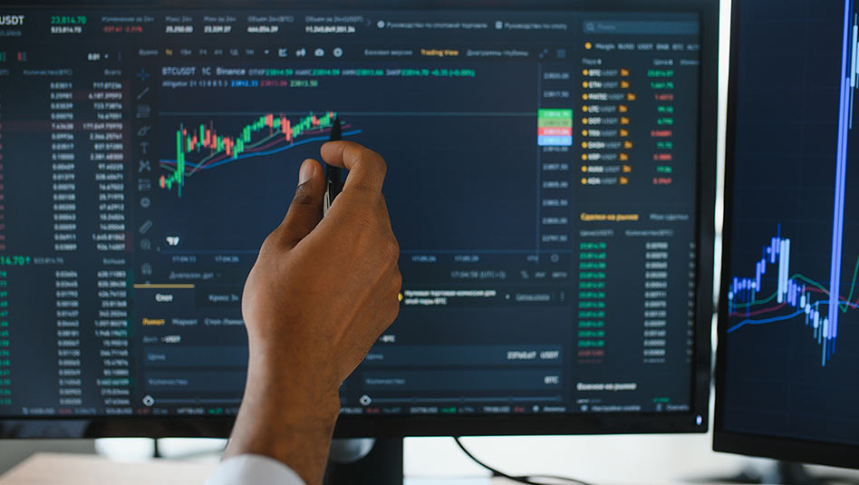 Hand holding a pen and pointing to a stock chart on a computer screen.