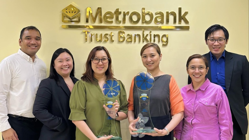 Four women and two men standing in front of a signage that says Metrobank Trust Banking. The two women in the middle are holding glass trophies.